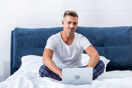 微笑的男性自由职业者看着相机和坐在床上与笔记本电脑在家里