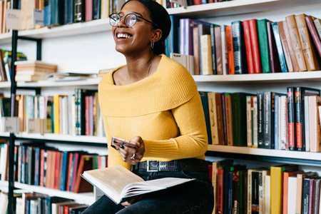 积极的非洲裔美国年轻女子在时尚眼镜视觉矫正笑, 而收到有趣的通知现代智能手机设备坐在现代图书馆的文学书籍
