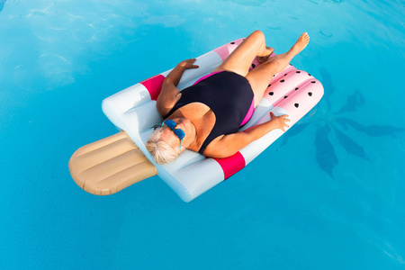 一位戴着明亮太阳眼镜的资深女性女子躺在游泳池充气冰淇淋形状的浮动享受阳光