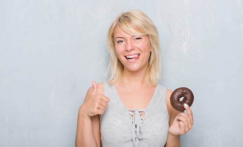 白人成年妇女在灰色的垃圾墙上吃巧克力甜甜圈快乐的大微笑做 ok 标志, 拇指与手指, 优秀的标志