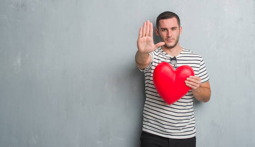 年轻的白种人在灰色的垃圾墙壁举行红色心脏用张开的手做停止标志以严肃和自信的表示, 防御手势