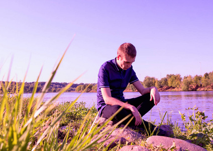 一个悲伤的年轻瘦家伙穿着蓝色衬衫, 坐在河边的石头上, 向下看