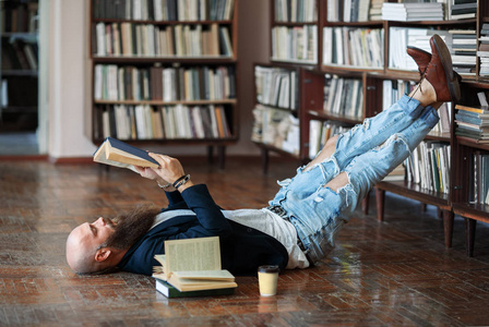 在图书馆躺在地板上的大胡子的时髦男人放松和读书书