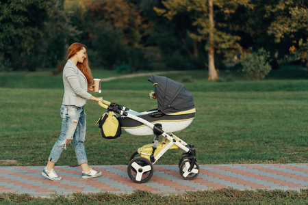 一个年轻的母亲喜欢公园散步在早上, 喝着热茶, 与她的小孩在他的婴儿推车放松