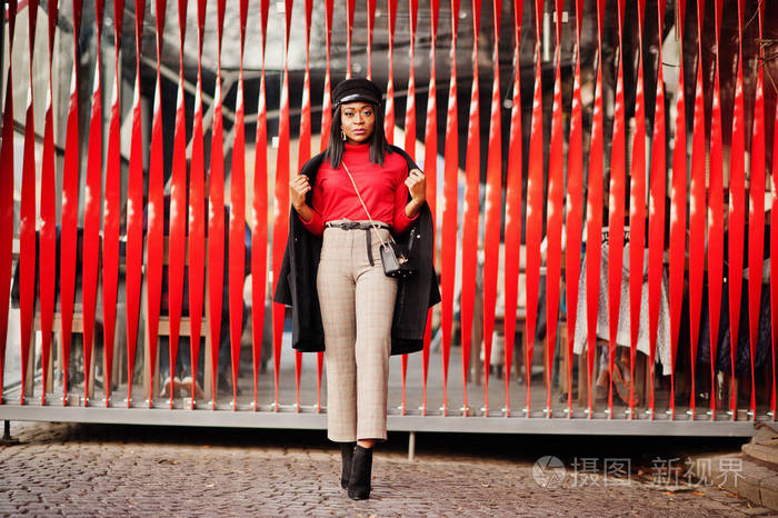 穿着外套和报童帽的非裔美国人时尚女孩在红色背景下在街上摆姿势