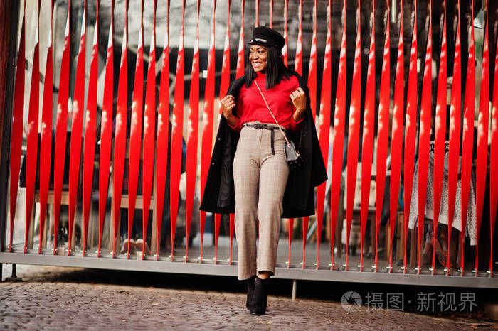 穿着外套和报童帽的非裔美国人时尚女孩在红色背景下在街上摆姿势