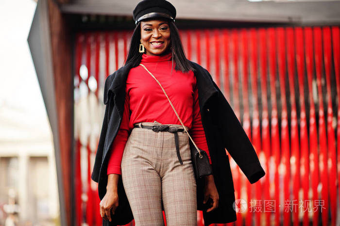 穿着外套和报童帽的非裔美国人时尚女孩在红色背景下在街上摆姿势