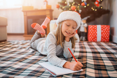 可爱的孩子躺在毯子上写信。她在下巴下又握着一只手, 两腿交叉。女孩一个人在房间里。她身后有圣诞树