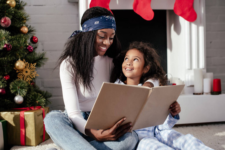 愉快的非洲裔美国母亲和女儿阅读书一起在家里, 圣诞节概念