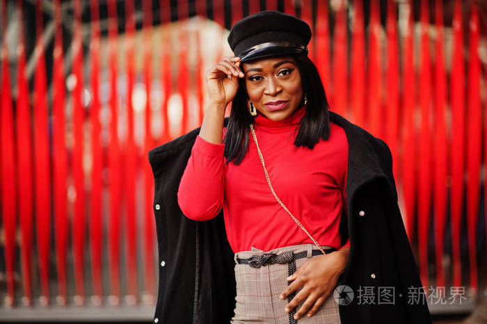 穿着外套和报童帽的非裔美国人时尚女孩在红色背景下在街上摆姿势
