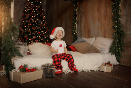 戴着圣诞老人帽子的可爱的小女孩正坐在房间里的圣诞树的背景上, 在一根棍子上吃着甜糖果