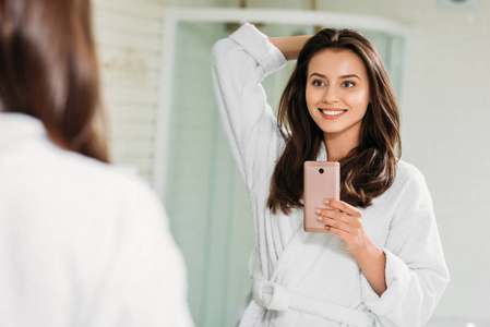 美丽的微笑的年轻妇女在浴袍采取自拍与智能手机在浴室里