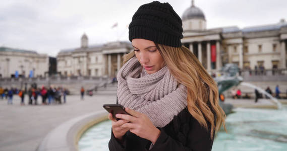 妇女在舒适的帽子和围巾短信与移动设备在特拉法加广场
