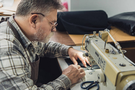 在工作室缝纫机上工作的围裙和眼镜的成熟男性裁缝的侧视图