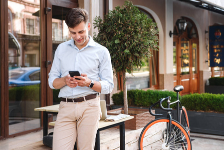 微笑的年轻时尚男子在衬衫上使用手机, 而站在一个咖啡馆户外与自行车