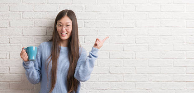 年轻的中国女人喝了一杯咖啡在白色砖墙非常高兴地指着手和手指的一侧