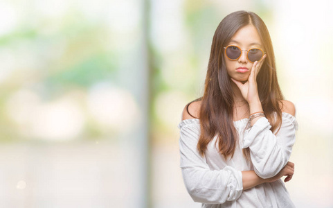 年轻的亚洲妇女戴着太阳镜在孤立的背景下思考疲倦和厌倦与交叉手臂的抑郁症问题