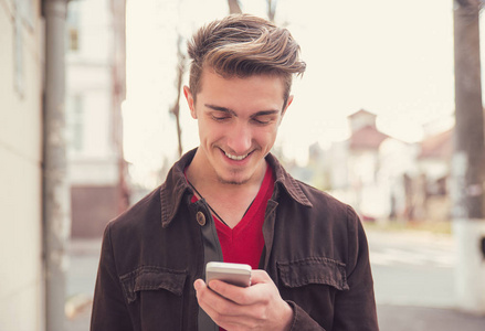 年轻男子在夹克使用智能手机, 而站在街道上, 愉快地微笑着