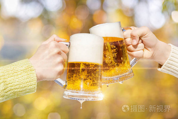 两只手是在模糊的秋季背景的新鲜泡沫啤酒杯