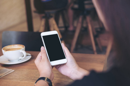 在餐厅的木桌上手持带咖啡杯的空白黑屏的手持白色手机的样机图像