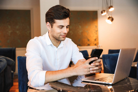 一个微笑的年轻正式穿衣男子的肖像手持手机工作在笔记本电脑上, 而坐在酒店大堂