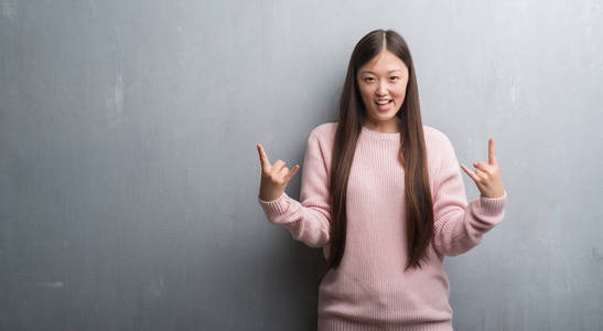 年轻的中国女人在灰色的墙上大喊着, 疯狂的表情举起手来做着摇滚符号。音乐明星。沉重的概念