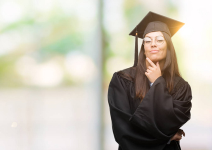 年轻的西班牙裔妇女穿着毕业帽和制服看着自信的相机与交叉的手臂和手举起下巴。积极思考