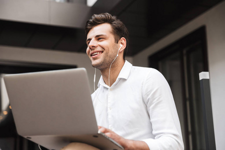 一个快乐的年轻正式穿着的人的肖像在笔记本电脑上工作, 而坐在户外的耳机