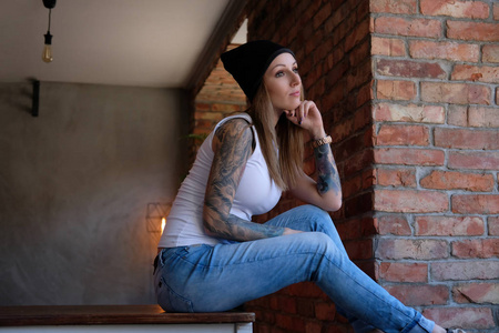 沉思的 tattoed 时髦的女孩穿着白色 t恤和帽子坐在桌子上, 看着窗外