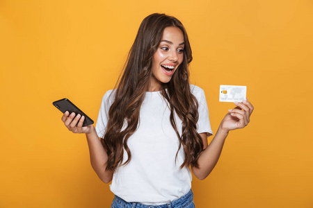 一个惊讶的年轻女孩与长黑发头发站立在黄色背景, 手持手机, 显示塑料信用卡的肖像