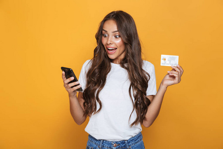 一个震惊的年轻女孩的肖像站在黄色背景, 手持手机, 显示塑料信用卡的长黑发头发