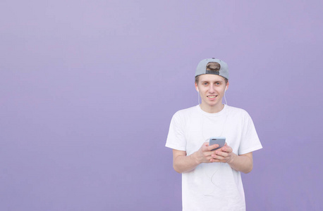 微笑的人站在他手中的智能手机在紫色柔和的背景, 听音乐的耳机和看相机。紫色墙背景下的一个年轻人的街道肖像