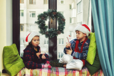 有趣的孩子们在等圣诞节。戴着圣诞老人帽子的兄妹在窗户上