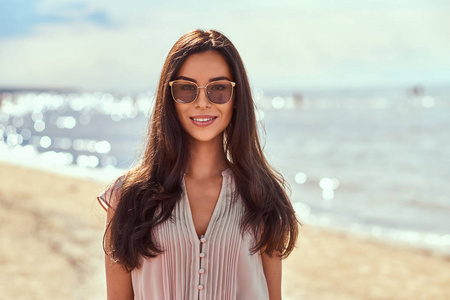 一个快乐的美丽的黑发女孩的肖像与长头发的太阳镜和礼服在海滩上