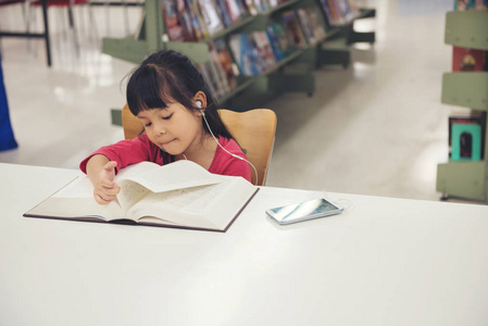 可爱的小女孩学习读书在图书馆室和书架背景书。儿童从智能手机上收听歌曲音乐