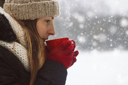 红色手套中的年轻女孩在降雪期间的冬季风景背景下举办了一个红色杯和热茶。冬季理念