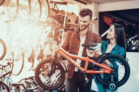 欢快的年轻夫妇选择儿童自行车在商店。美丽的女孩站在体育自行车店看帅哥穿着格子衬衫抱着孩子的自行车在手和笑