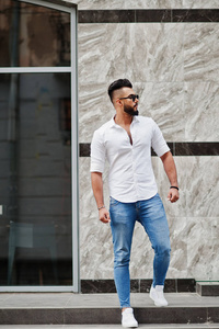 时尚高大的阿拉伯男子模特在白色衬衫, 牛仔裤和太阳镜摆在城市的街道。胡子迷人的阿拉伯家伙