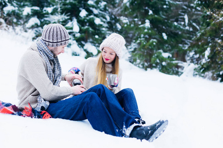 一对夫妇戴着帽子和毛衣坐在雪地里, 那个家伙从热水瓶里倒了一杯热饮, 抱着女孩