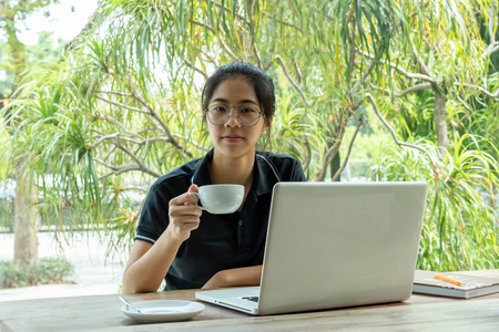 美丽的年轻亚洲女孩拿着咖啡与笔记本电脑在咖啡馆的桌子上