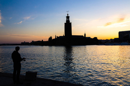 在瑞典, 在日落时分, 黄昏, 暮色中, 在孔舒门岛上, 渔民用钓竿和斯德哥尔摩市政厅 stadshuset 建造市议会的剪影