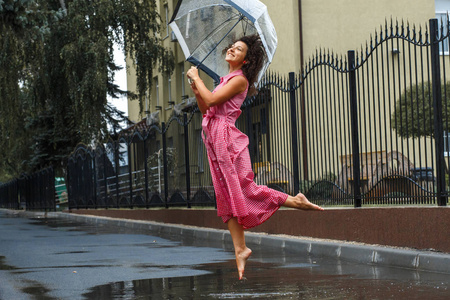 年轻的女孩穿着红色的连衣裙, 一把透明的雨伞, 在雨中跳舞, 站在水坑里