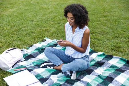 形象的愉快的微笑年轻的非洲女孩坐在草地上户外公园聊天通过电话