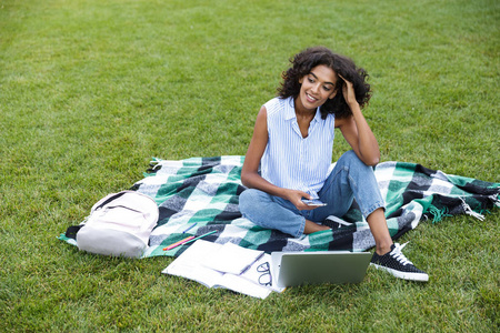 形象的愉快的微笑年轻的非洲女孩坐在草地上户外公园聊天通过电话