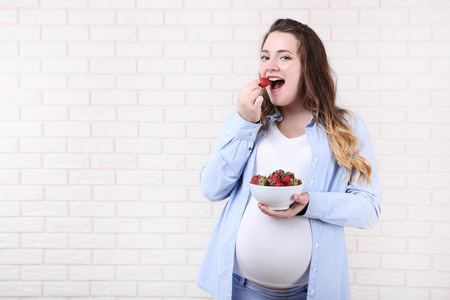 美丽的怀孕妇女与草莓在碗在砖墙背景