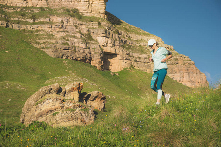 有吸引力的运动女孩在帽子和耳机慢跑在一个风景如画的地方下山附近的岩石在日落。户外锻炼