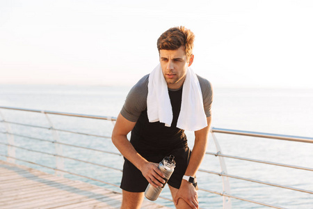 成人运动员的相片20s 在运动服与毛巾在脖子饮用水从热水瓶在海滩的木码头在早晨锻炼以后
