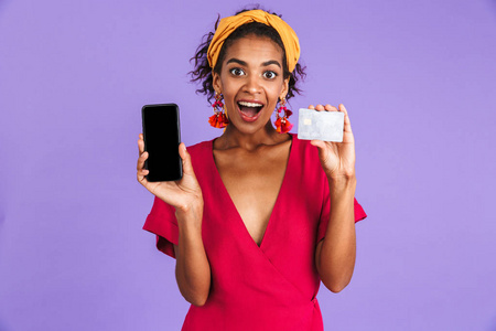 开朗的非洲妇女在服装持有信用卡和显示空白智能手机屏幕, 而看着相机上空紫色背景