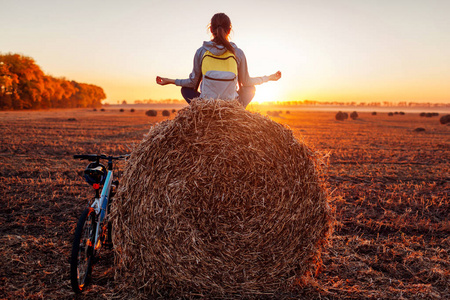 年轻的自行车冥想坐在干草堆后骑。在秋季领域休息练习瑜伽的妇女。运动娱乐概念