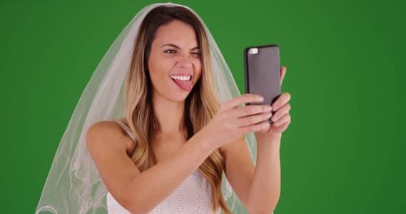 年轻的新娘在婚纱采取有趣的自拍与手机在绿色屏幕上
